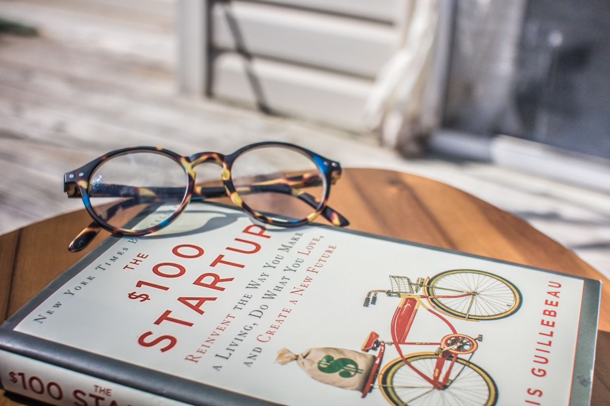 A pair of reading glasses sit atop the book The $100 Startup, which sits on a small table on a patio.